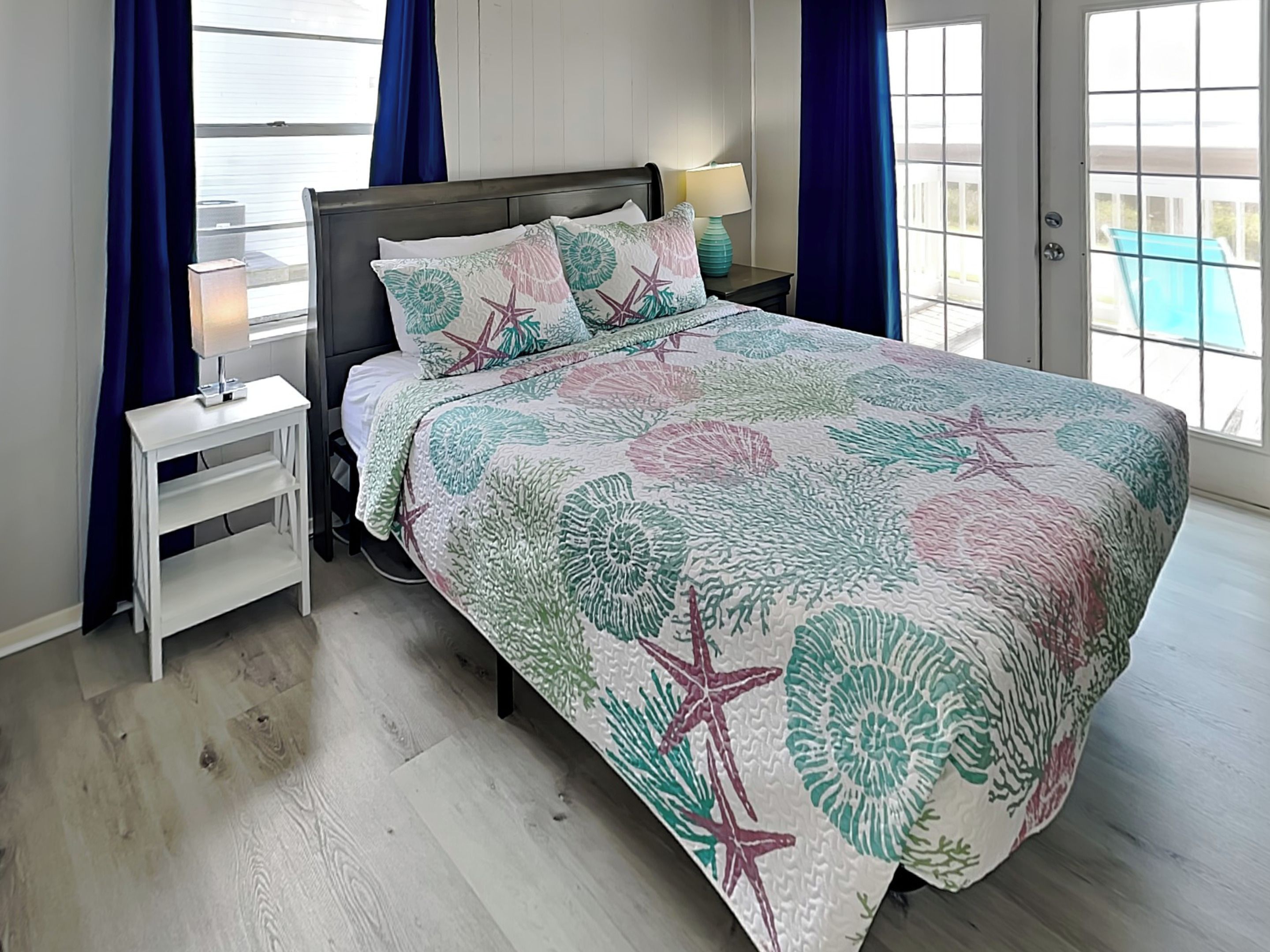 A bedroom with a bed, nightstands, a lamp, and a view of the outdoors.