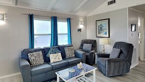 A living room with a grey sofa, a matching armchair, a small white coffee table, and a painting on the wall. - Bayview Cottage by Ryson Vacation Rentals (Galveston)