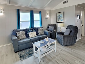 A living room with a grey sofa, a matching armchair, a small white coffee table, and a painting on the wall. - Bayview Cottage by Ryson Vacation Rentals (Galveston)