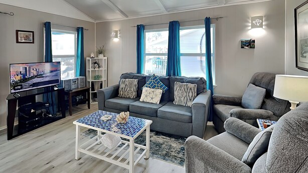 A modern living room with a grey sofa, a coffee table, and a TV. There are blue curtains, a white lamp, and a framed picture on the wall. - Bayview Cottage by Ryson Vacation Rentals (Galveston)