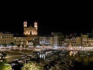 Vista noturna de um edifício histórico com duas torres, uma área à beira-mar com barcos e uma rua movimentada com lojas e pessoas.
