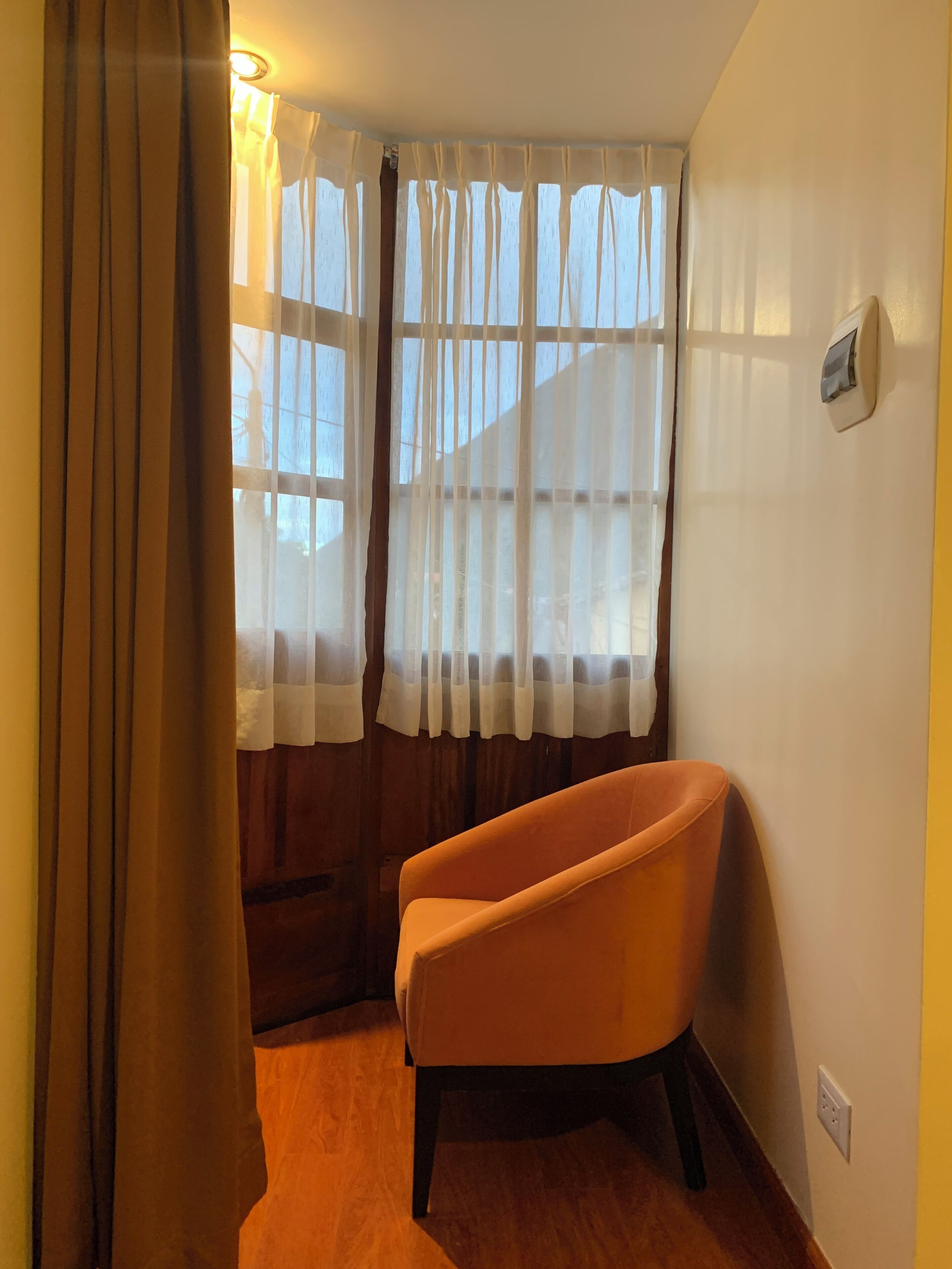 A room with a wooden floor, a single orange armchair, a window with sheer curtains, and a wall-mounted thermostat.