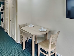 Un pequeño comedor con una mesa redonda y dos sillas, una taza blanca sobre la mesa, y una puerta con manija al fondo.