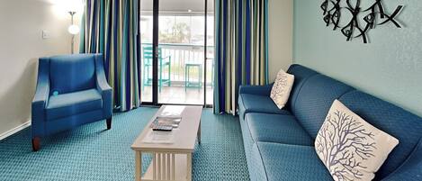 Une pièce avec un canapé bleu, un fauteuil bleu, une petite table en bois et, à travers une porte vitrée, une vue sur un espace extérieur aménagé avec des chaises.