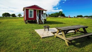 A red wooden cabin with a porch and a picnic table in a grassy field.