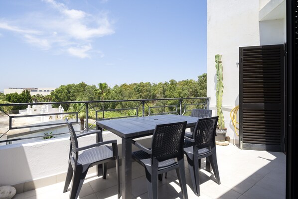 A balcony with a table and chairs, a potted plant, and a view of a building and trees.