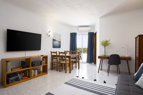 A modern living room with a flat-screen TV, wooden bookshelves, a dining area with chairs, a desk with a chair, and a sofa. - Albufeira Salgados Sailor by Hopstays | Balcony (Albufeira)