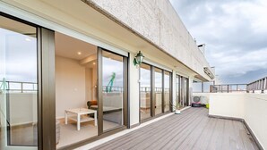 A modern balcony with a wooden floor, large sliding glass doors leading to an interior space, and a view of a bridge in the background.