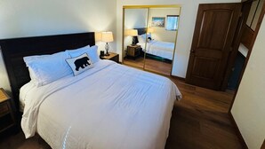 A hotel room with a large bed, a nightstand with a lamp, a wooden door, and a mirror reflecting the room. - Walk to Village - 2 Bdrm, +Loft, 2 Bath - Mammoth Haus (Mammoth Lakes)