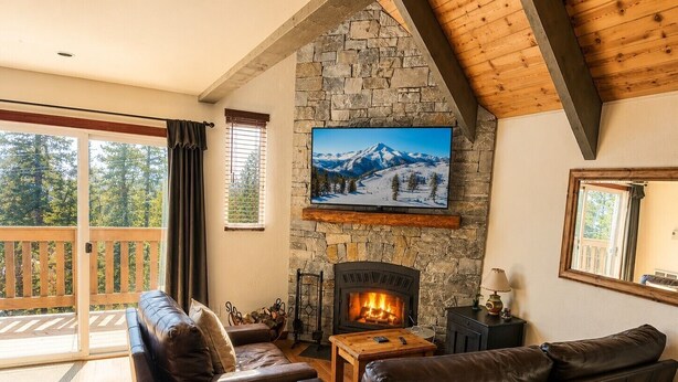 A cozy living room with a stone fireplace, leather sofas, a wooden coffee table, and a large window overlooking a deck. - Walk to Village - 2 Bdrm, +Loft, 2 Bath - Mammoth Haus (Mammoth Lakes)