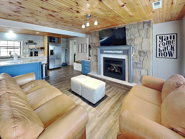 A modern living room with leather sofas, a stone fireplace, and a kitchen in the background. - New Era Peak by Jackson Mountain Rentals (Sevierville)