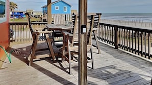 A wooden deck with a table and chairs, overlooking a beach and a blue house in the background. - Once Upon a Tide by Ryson Vacation Rentals (Galveston)