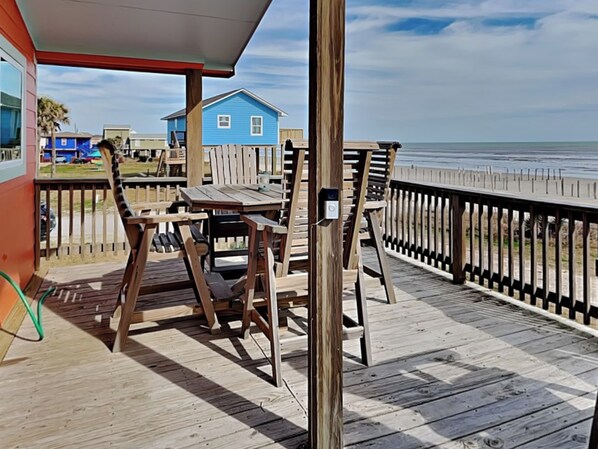 A wooden deck with a table and chairs, overlooking a beach and a blue house in the background.