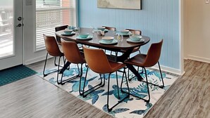 A dining area with a round table set for six, surrounded by six brown chairs. - Once Upon a Tide by Ryson Vacation Rentals (Galveston)