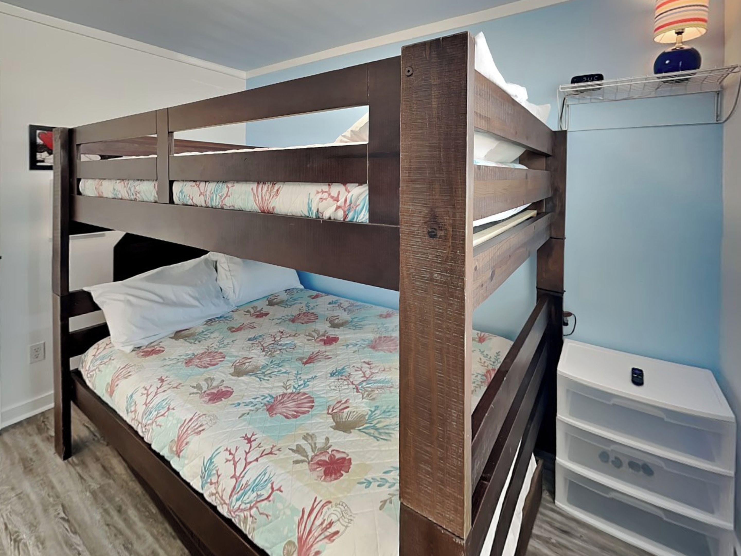 Un lit superposé avec une couvre-lit à motifs floraux, une table de chevet avec une lampe et un meuble de rangement blanc.