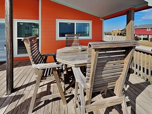 A wooden deck with a table and chairs, a railing, and a building with orange siding and white trim in the background. - Once Upon a Tide by Ryson Vacation Rentals (Galveston)