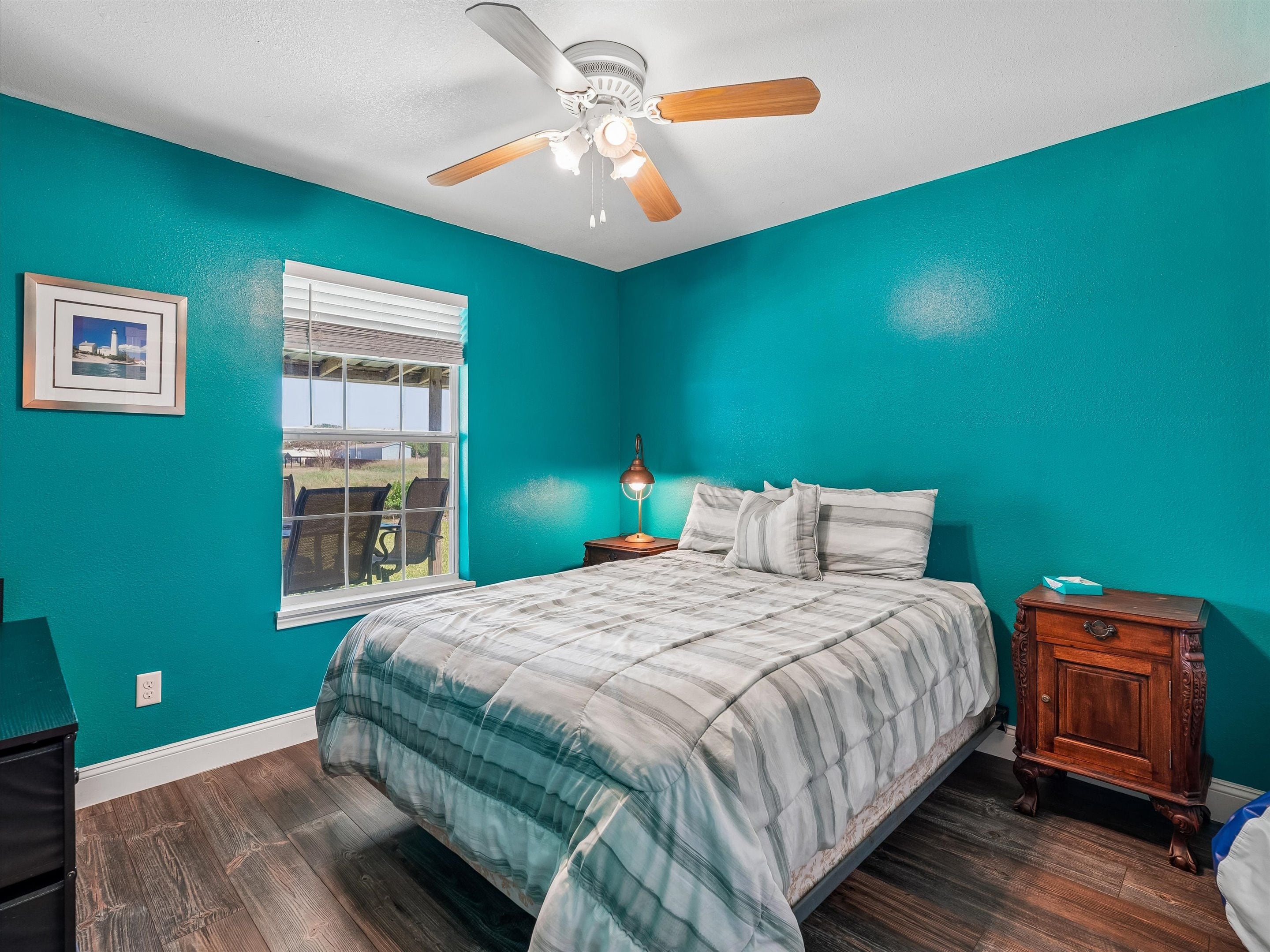 A bedroom with teal walls, a bed with a striped comforter, a ceiling fan, a bedside table with a lamp, and a window with blinds.