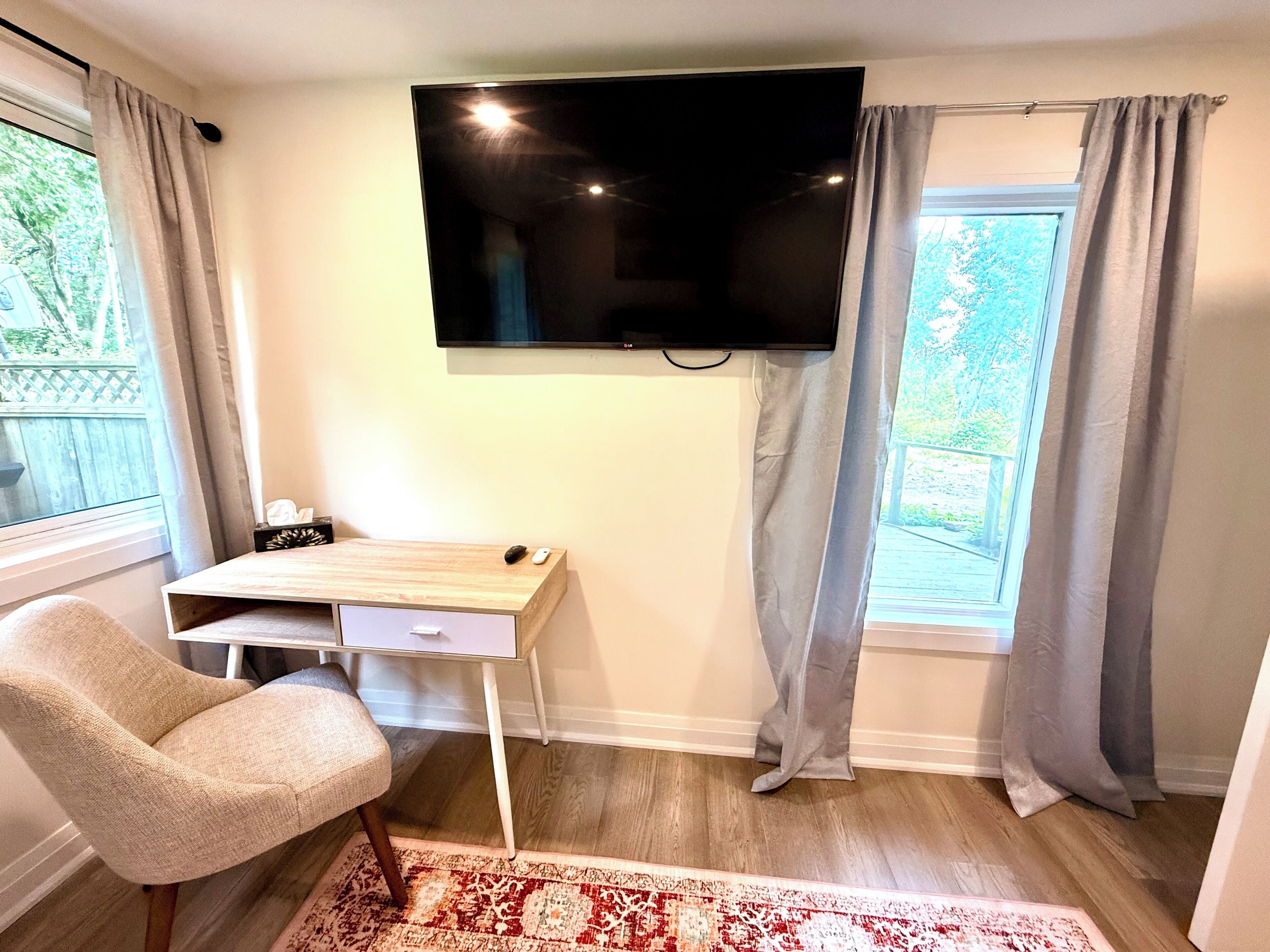 Une pièce dotée d’un téléviseur à écran plat fixé au mur, d’un bureau en bois avec un ordinateur, d’une chaise et d’une fenêtre avec des rideaux.