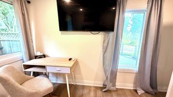 Une pièce dotée d’un téléviseur à écran plat fixé au mur, d’un bureau en bois avec un ordinateur, d’une chaise et d’une fenêtre avec des rideaux.