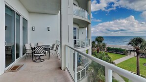 A balcony with a table and chairs, a view of the ocean, and a well-maintained lawn with palm trees. - Low Tides by Ryson Vacation Rentals (Galveston)