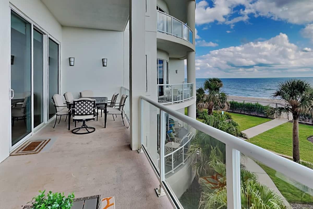 A balcony with a table and chairs, a view of the ocean, and a well-maintained lawn with palm trees.