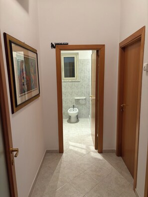 Un couloir mène à une salle de bain équipée d’un bidet. Un tableau encadré est accroché au mur, et une pancarte au-dessus de la porte indique : « SALLE DE RÉFLEXION ».