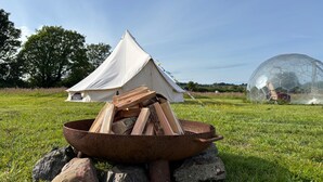 Ein Zelt, eine Geodätkuppel und eine Feuerstelle mit Holzscheiten in einer grünen Wiese.