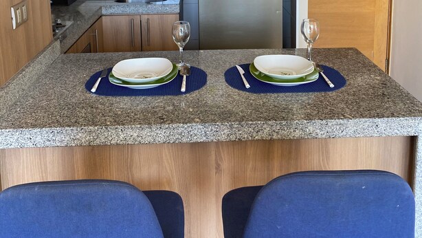 Una cocina compacta con armarios de madera, un refrigerador de acero inoxidable, un microondas y un lavamanos. Hay dos zonas de comedor con individuales azules y copas de vino.