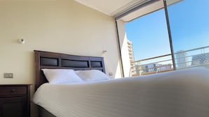 Un dormitorio con una cama grande, una mesita de noche, una lámpara de pared y una ventana con vistas a edificios.
