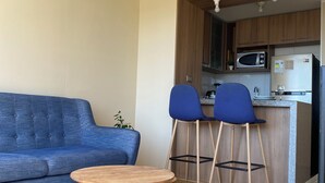 Un espacio habitable pequeño con un sofá, dos taburetes, una mesita redonda y una kitchenette con microondas y refrigerador.