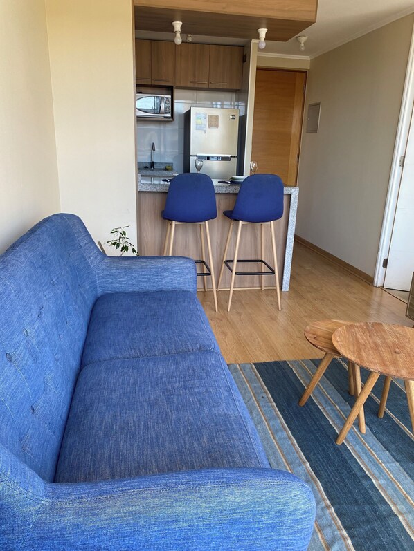 Una sala moderna con un sofá azul, dos sillas azules, una pequeña mesa de madera y una zona de cocina al fondo.