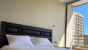 Un cuarto de hotel con una cama grande, un cabecero oscuro y un ventanal con vista a un edificio alto.