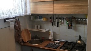 Eine kleine Küche mit Holzausziehtisch, einem Gasherd, einer Spüle und einem Fenster mit weißem Vorhang.