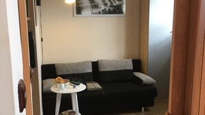 Ein gemütliches Wohnzimmer mit einer schwarzen Couch, einem kleinen runden Tisch mit einer Lampe, einer Uhr an der Wand und einem gerahmten Bild über der Couch.