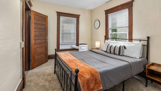 A bedroom with a bed, two windows with blinds, a wooden door, a nightstand, and a wall clock. - HIstoric 4 BR Near Fulton St (Grand Rapids)