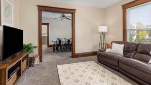 A living room with a brown leather sofa, a TV on a wooden stand, a patterned rug, and a dining area in the background. - HIstoric 4 BR Near Fulton St (Grand Rapids)