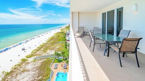 Un balcón con una mesa y sillas de vidrio, con vista a una playa que tiene una piscina y sombrillas.