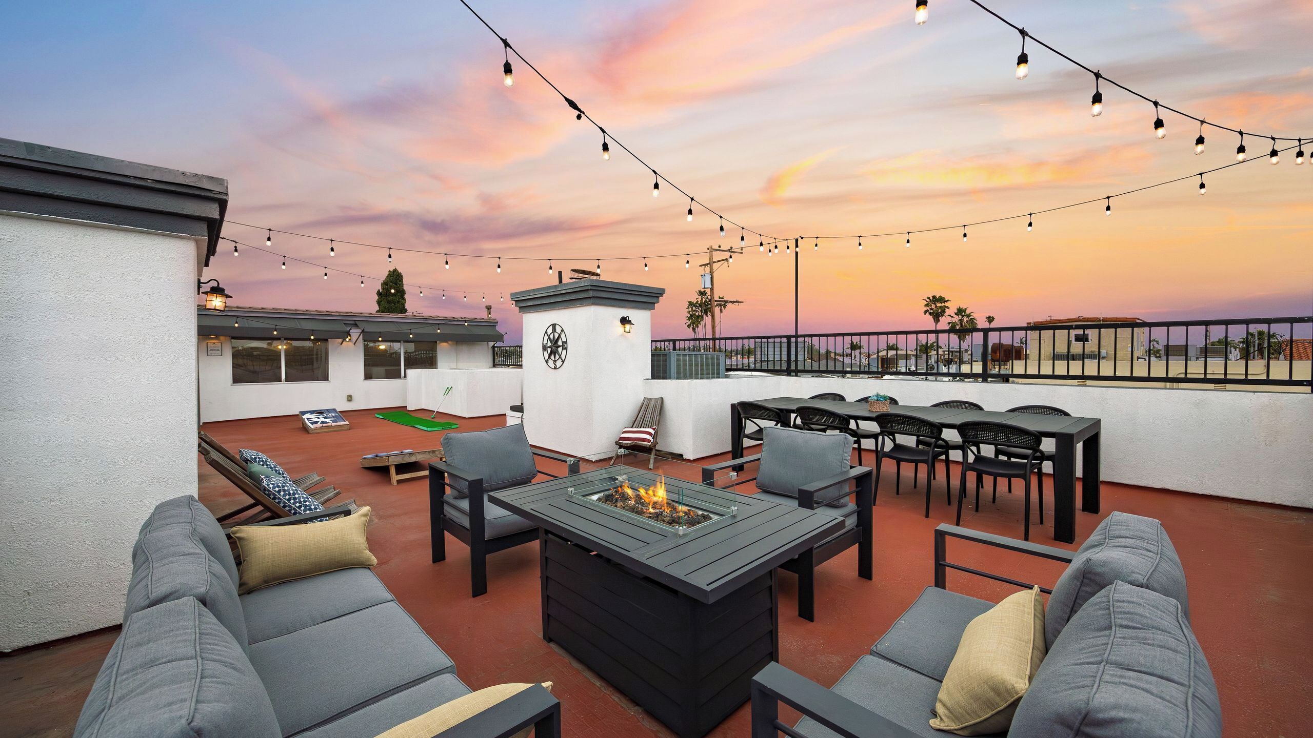 An outdoor patio with seating, a fire pit, string lights, and a view of the sunset.