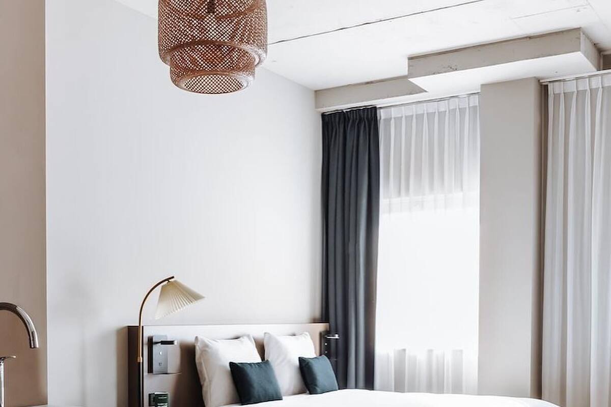 A modern hotel room with a bed, a desk with a lamp, a window with curtains, and a wooden floor.