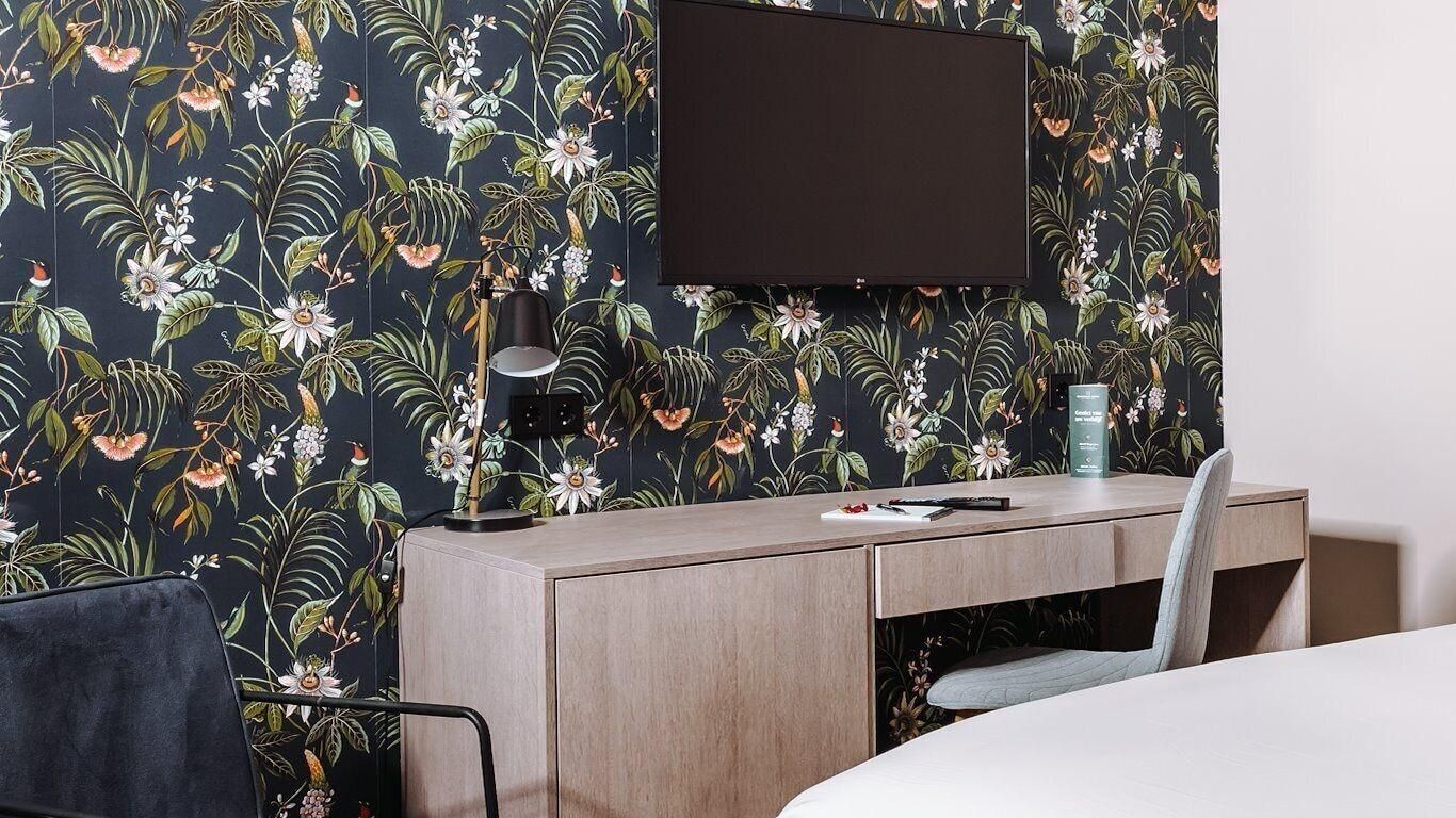 A hotel room with a bed, a desk, a chair, a TV mounted on the wall, and a floral wallpaper.