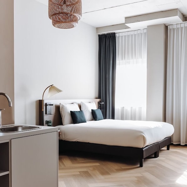 A modern hotel room with a bed, a desk with a lamp, a window with curtains, and a wooden floor.