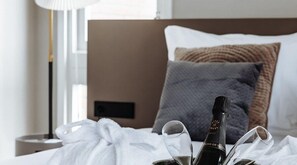 A neatly made bed with a bottle of champagne and glasses, a bedside lamp, and a window with curtains. - Havendijk Hotel (Schiedam)