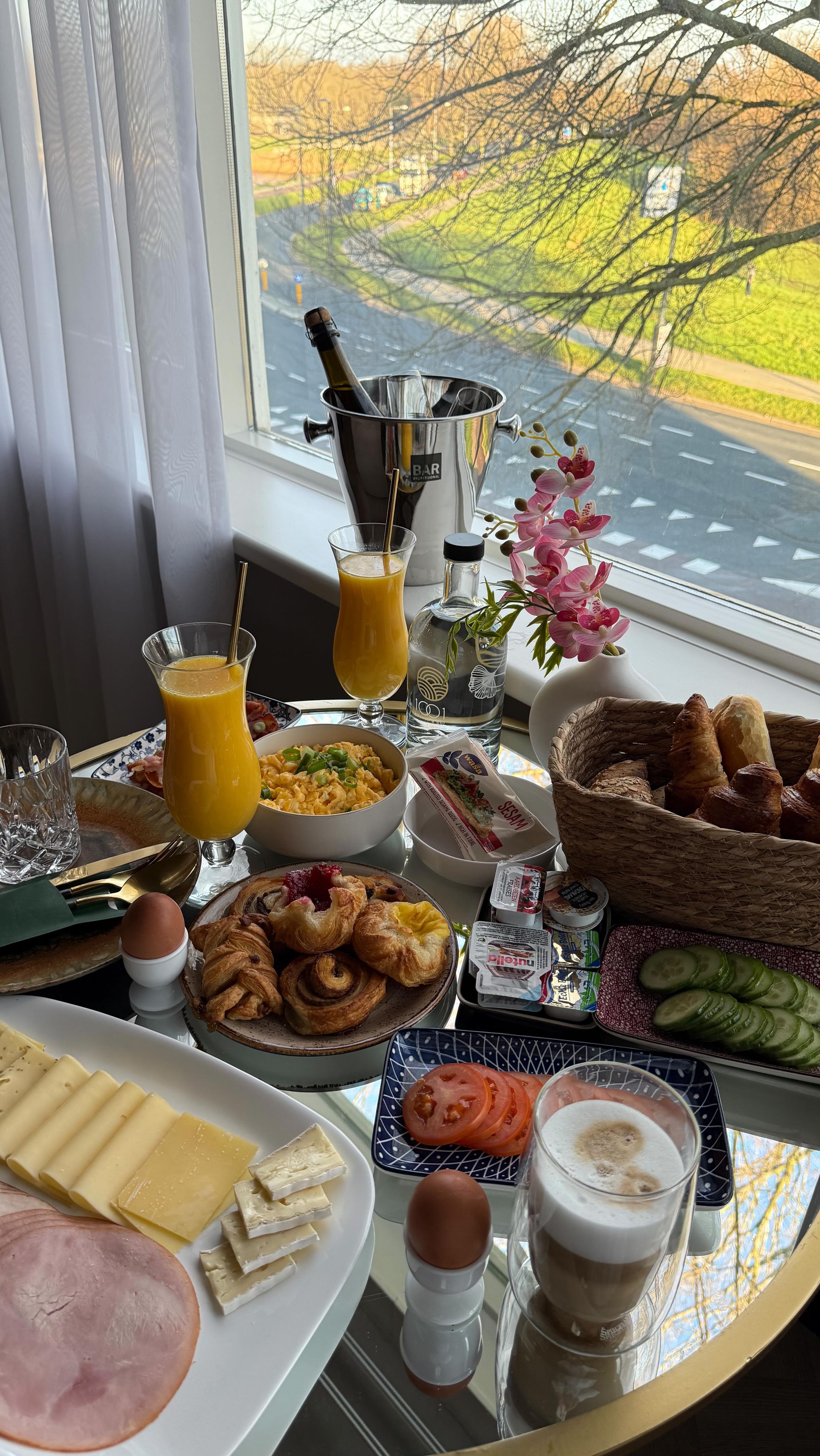Une table de petit-déjeuner avec divers aliments, une bouteille de champagne et une vue sur une route et des arbres, visible à travers une fenêtre.