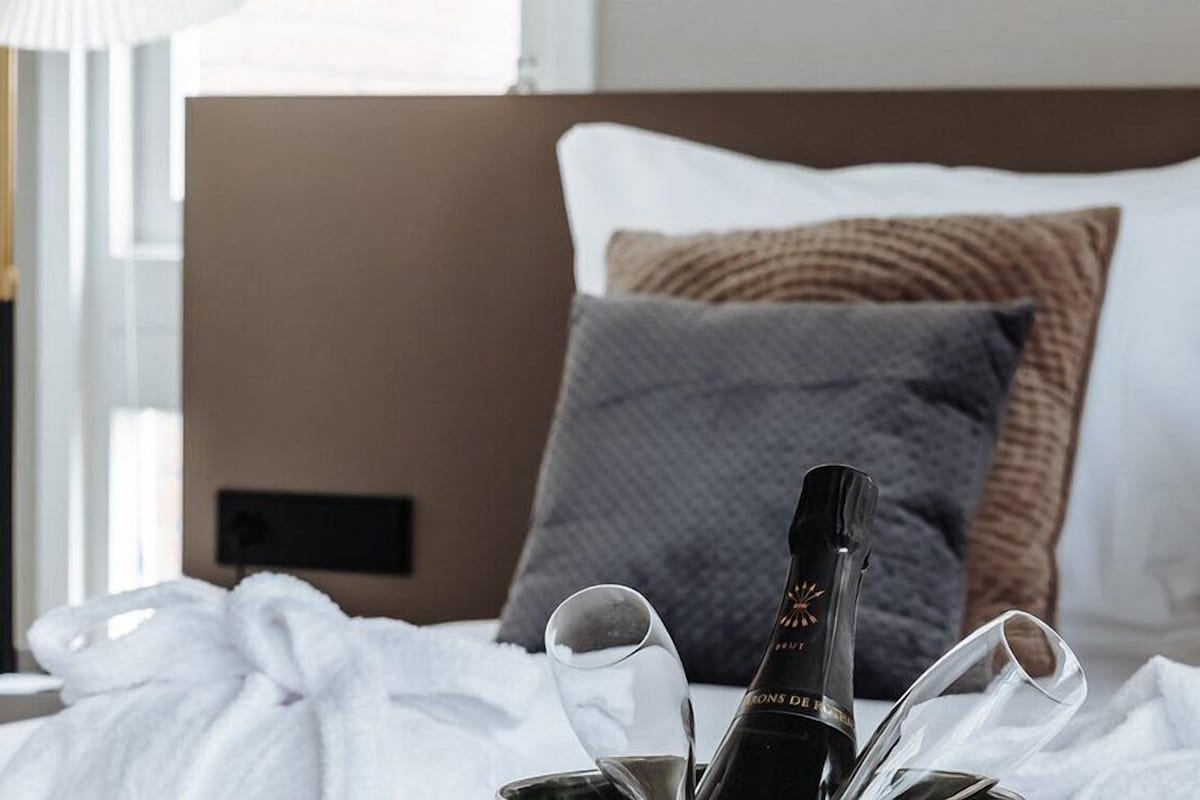 A neatly made bed with a bottle of champagne and glasses, a bedside lamp, and a window with curtains.