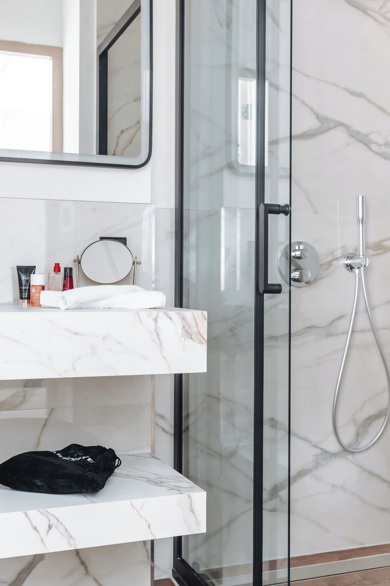 Un baño con puerta de vidrio para la ducha, un lavabo de mármol blanco y una bolsa negra sobre el mostrador.