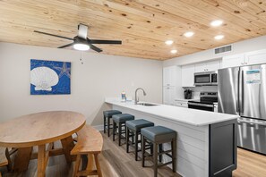 A modern kitchen with a wooden ceiling, a central island with stools, a stainless steel refrigerator, and a microwave. - Blue Surf 22 (Miramar Beach)