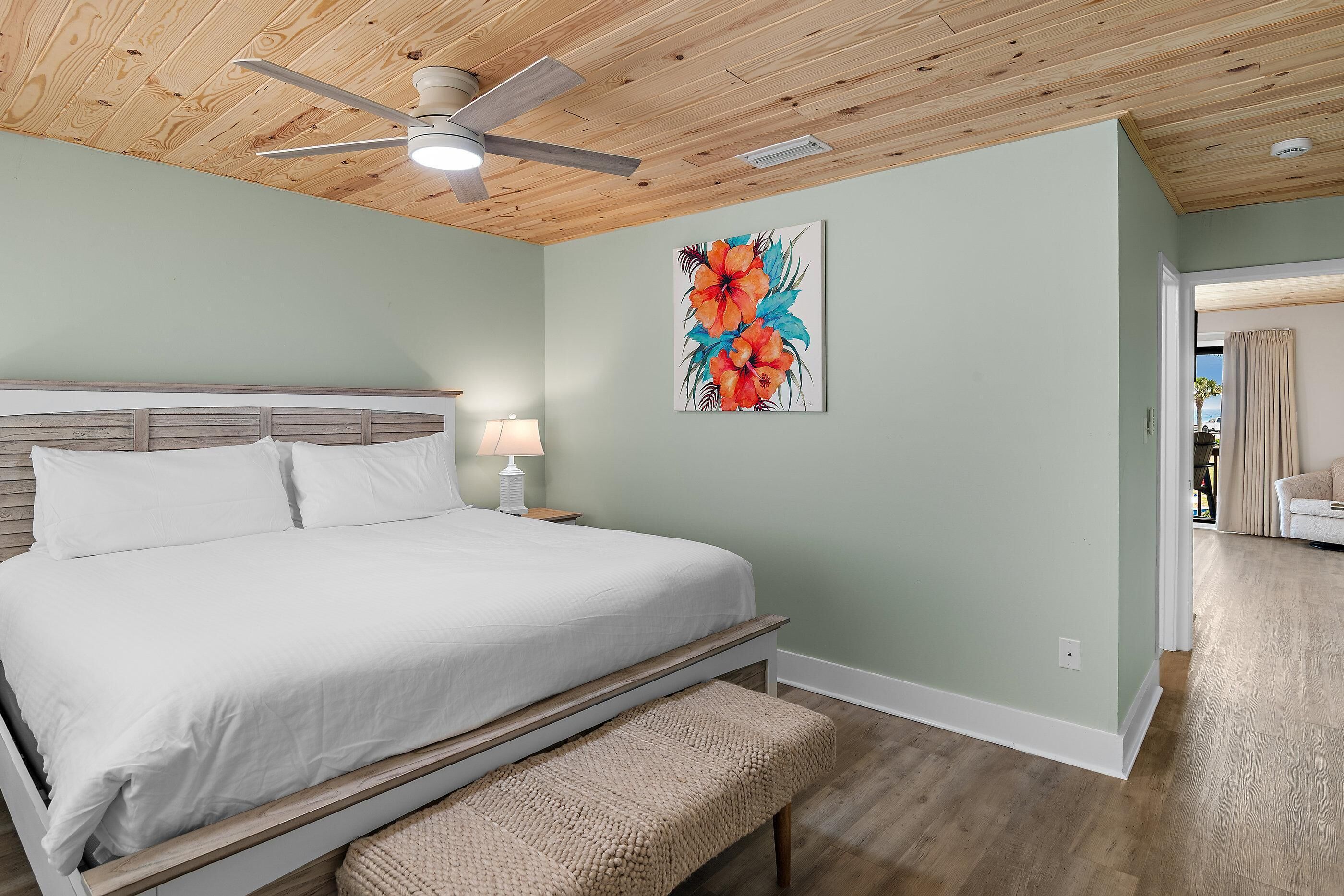 Un dormitorio con una cama, un ventilador de techo, una mesita de noche con lámpara y una pared decorada con un cuadro floral.