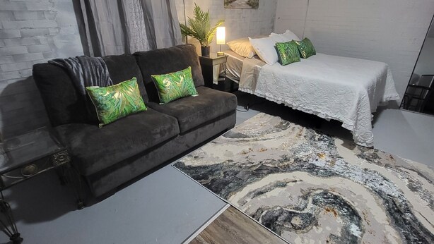 A modern bedroom with a grey brick wall, a grey sofa with green cushions, a bed with white bedding, and a wooden floor. - Charming 1-bedroom apartment in amazing Wichita with WiFi, AC (Wichita)