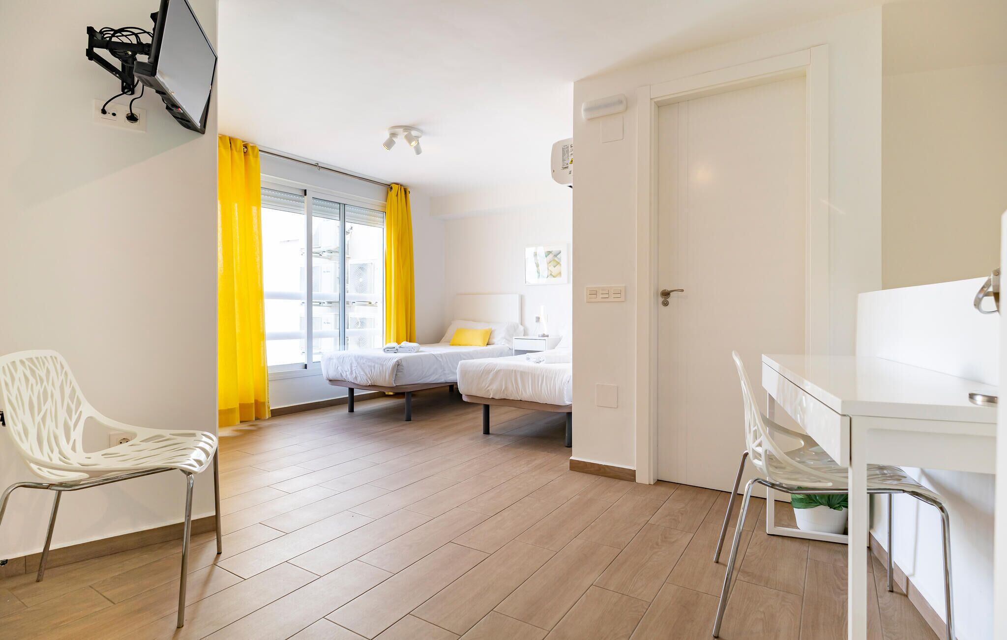 A modern hotel room with two beds, a desk, a chair, and a window with yellow curtains.