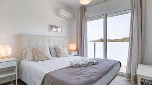 Une chambre moderne avec un grand lit, du linge de lit blanc, une table de chevet avec une lampe, et une vue sur la mer grâce à une porte-fenêtre coulissante.